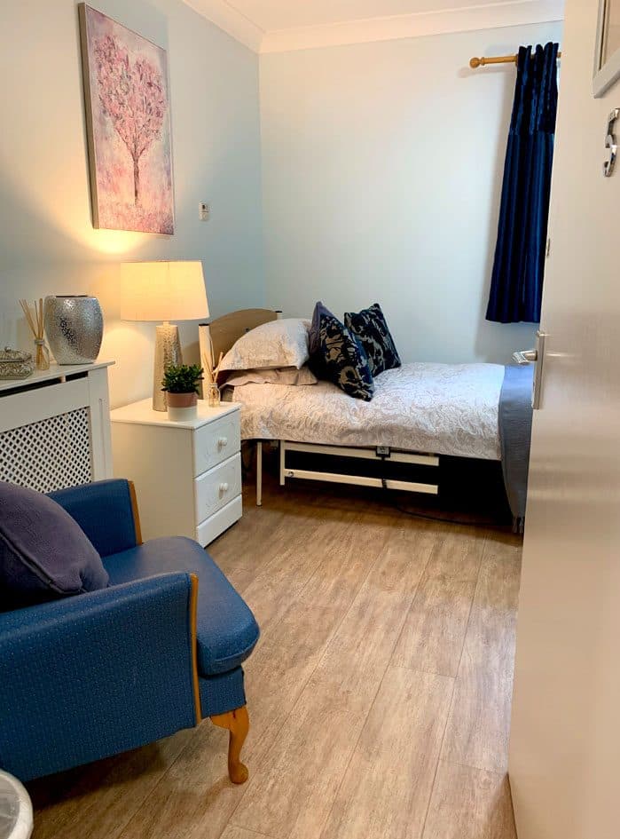 A bedroom at Grafton Lodge with a made bed, a lamp, a painting and a blue armchair